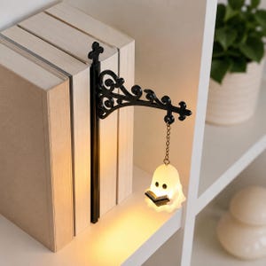 May include: A black metal bookend featuring an ornate, scrollwork bracket arm that extends outward. A small, illuminated white ghost figure holding a tiny black book hangs from the end of the arm by a short metal chain, casting a warm glow.