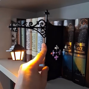 May include: A black metal bookend featuring an ornate, scrollwork arm that extends to hold a miniature, glowing lantern suspended by a small chain. The structure includes a decorative fleur-de-lis emblem on the side panel, positioned between books.