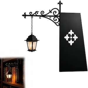 May include: A black metal wall-mounted sign bracket featuring an ornate scrollwork arm and a decorative fleur-de-lis cutout. A small, four-sided lantern with clear glass panels hangs from the bracket by a short metal chain, emitting a warm light.