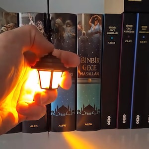 May include: A miniature black lantern emitting a warm, bright yellow light, held in front of a row of books titled Binbir Gece Masallari. The lantern features a classic hexagonal frame design with a small hanging loop at the top and a glowing center.