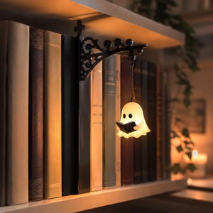 May include: A black metal decorative wall bracket with ornate scrollwork, featuring a small, glowing, cream-colored ghost figurine hanging from a chain. The ghost has two black eyes and is holding a small, open black book in its hands.