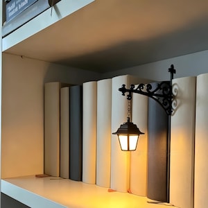 May include: A black metal wall-mounted lantern light fixture with an ornate scrollwork bracket, featuring a small hanging lamp with a warm glowing light, positioned between several books on a shelf to serve as a decorative bookend accessory.