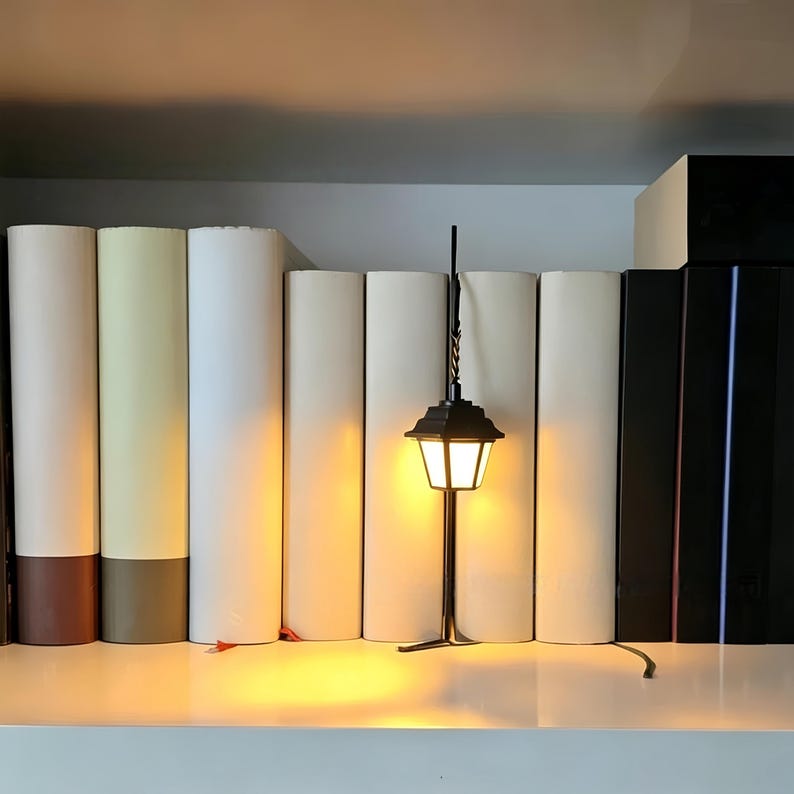 May include: A small, black, metal lantern-style lamp with a warm, glowing light, featuring a tapered glass shade and a thin, vertical support post, positioned in front of a row of solid-colored, neutral-toned books on a shelf.