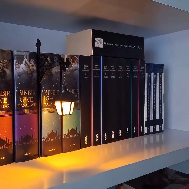 May include: A black metal street lamp miniature with a warm glowing light, positioned between two books titled Binbir Gece Masallari. The lamp features a classic lantern design and is placed on a shelf next to a collection of black books.