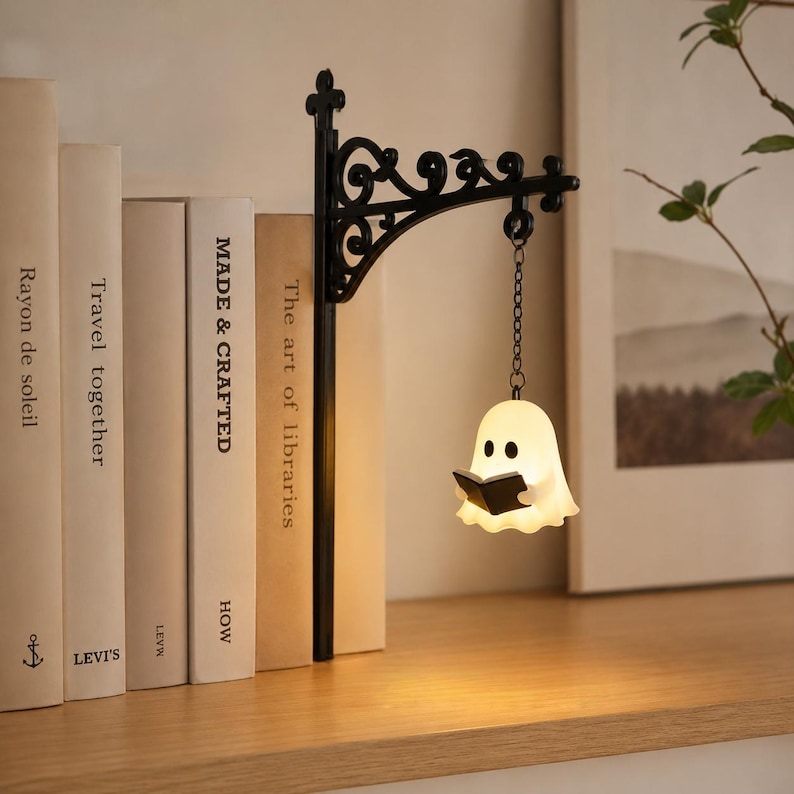 May include: A black metal wall bracket with ornate scrollwork supports a small, glowing white ghost figurine hanging from a chain. The ghost has black eyes and holds a small black book. The lamp emits a soft light, illuminating the surrounding area.