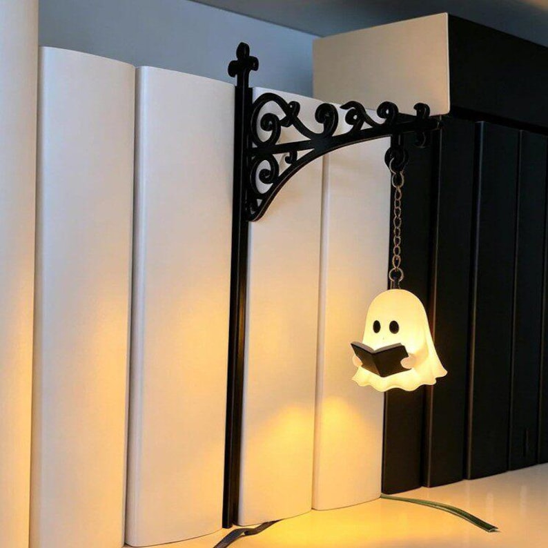 May include: A black metal wall-mount style bracket with ornate scrollwork, featuring a small, glowing white ghost figurine holding a black book. The ghost is suspended from the bracket by a silver-toned metal chain, creating a decorative bookend.