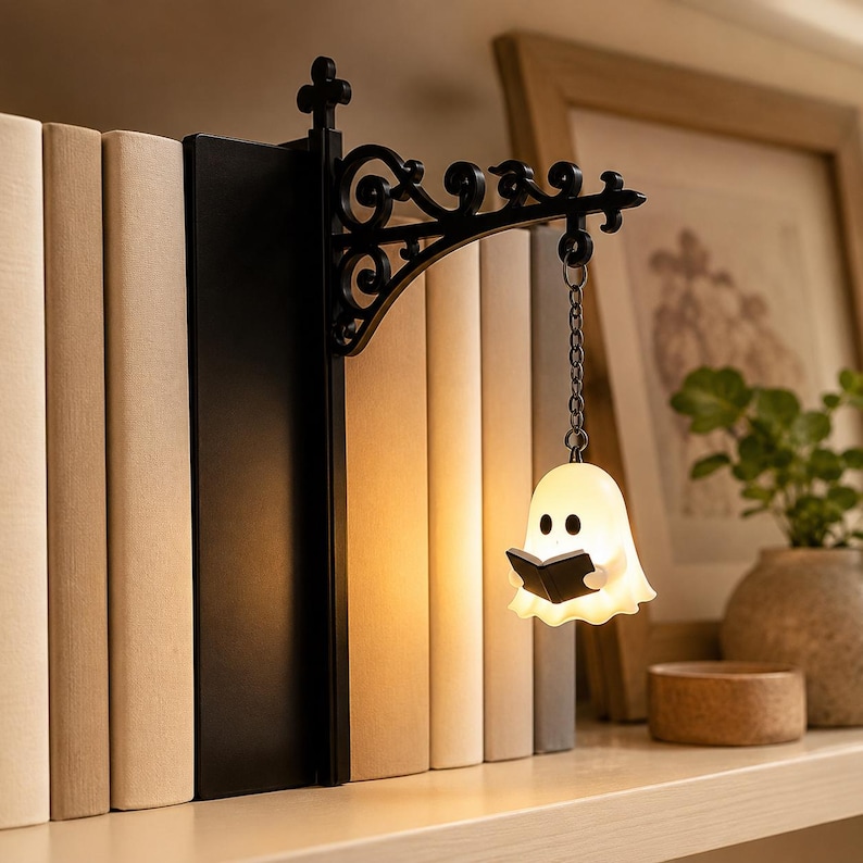 May include: A black metal bookend featuring an ornate, scroll-work bracket that extends outward. A small, white, glowing ghost figure holding a black book hangs from the bracket by a metal chain, serving as a light source against a row of books.
