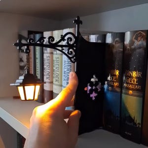 May include: A black metal bookend featuring an ornate, scrollwork arm that extends to hold a miniature, glowing lantern suspended by a small chain. The structure includes a decorative fleur-de-lis emblem on the side panel, positioned between books.