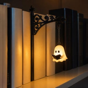 May include: A black metal bookend featuring an ornate, scrollwork bracket that extends outward. A small, glowing, white ghost figure holding a black book hangs from the bracket by a silver-toned metal chain, emitting a warm, soft light.