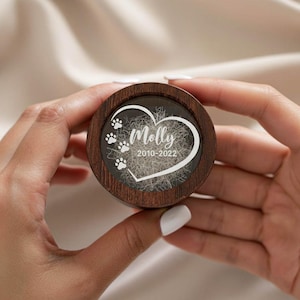Puede incluir: Una caja de recuerdos redonda de madera con tapa transparente. La tapa presenta un diseño en forma de corazón con el nombre "Molly", huellas de patas y los años "2010-2022". La caja está sujeta con dos manos.
