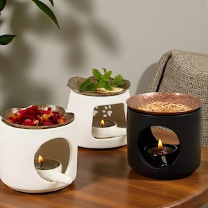 May include: Three ceramic oil warmers in white and black, each with a metal dish on top. One warmer holds rose petals, another mint and lemon slices, and the third, dried herbs. Each warmer has a lit tea light candle inside.