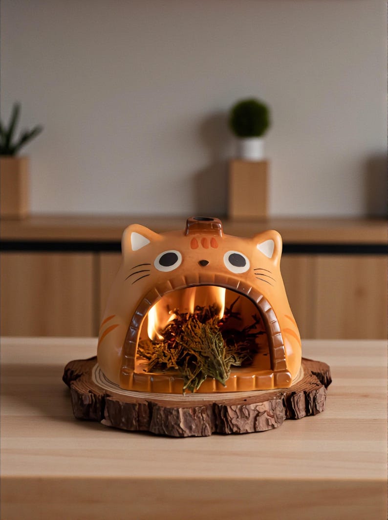 May include: A ceramic cat-shaped incense burner in shades of orange and brown, with a small chimney and a large opening where incense is burning. The burner sits on a round wooden base, creating a cozy, decorative piece.