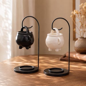 May include: Two ceramic cauldron-shaped wax warmers, one black and one white, each suspended from a black metal stand with a circular base. The small pots feature three feet and a handle, hanging from a curved hook designed for heating wax melts.