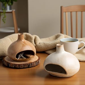 Op de afbeelding: Twee bolvormige keramische wierookbranders, één lichtbruin op een houten schijf, de andere gebroken wit. Beide hebben een opening aan de zijkant. Een mok thee en een beige deken staan op de achtergrond.