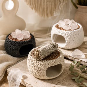 May include: Three round, textured stone oil burners with copper-colored metal mesh tops. One burner is black, two are off-white. The black burner holds pink quartz crystals, one white burner holds clear crystals, and the other holds a sage bundle.