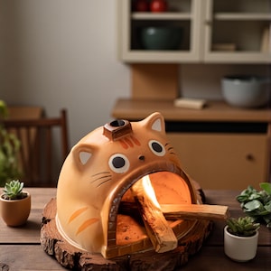 Op de afbeelding: Een keramische katvormige wierookbrander in oranje en bruine tinten, met een kleine schoorsteen en een brandende wierookstok aan de binnenkant. De brander staat op een houten voet, met kleine potplanten aan weerszijden. De achtergrond omvat een houten tafel en kasten.