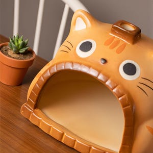 May include: A ceramic cat-shaped planter in shades of orange and brown. The planter has a large opening for plants, with black eyes and whiskers. A small succulent plant in a terracotta pot sits nearby. The cat planter is a decorative item.