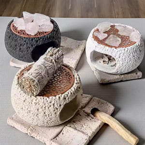 May include: Three decorative stone bowls with a textured surface. One is black, and two are white. Each bowl has a copper-colored grate and is filled with crystals or a bundle of sage. The bowls sit on stone tiles.