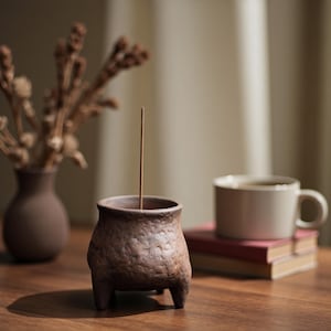 Puede incluir: Un porta incienso de cerámica marrón con tres patas pequeñas, que sostiene una sola varilla de incienso. Un jarrón con flores secas y una taza de té sobre una pila de libros están en el fondo. Los objetos están sobre una superficie de madera.
