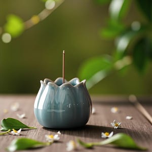 Pode incluir: Um porta-incenso de cerâmica azul claro em forma de flor de lótus, com um único incenso. O suporte está sobre uma superfície de madeira, rodeado por pequenas flores brancas e folhas verdes. O fundo é verde suave.