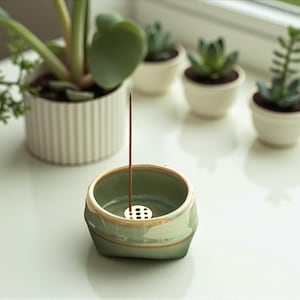 May include: A light green ceramic incense holder with a gold rim, holding a brown incense stick. The holder is on a white surface, with small potted succulents in the background. The holder has a small, perforated metal insert.