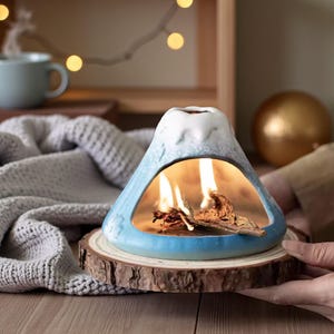 Puede incluir: Un difusor de aceites esenciales de cerámica con forma de volcán, en tonos azules y blancos, sobre una base de madera. El difusor tiene una llama en su interior, creando un ambiente cálido. El producto está diseñado para aromaterapia.