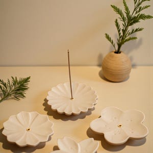 Könnte beinhalten: Eine Sammlung cremefarbener Keramik-Räucherstäbchenhalter in verschiedenen Blütenformen. Ein Halter hat einen Räucherstäbchen. Eine kleine Holzvase mit Grünzeug befindet sich im Hintergrund. Die Halter haben ein kleines goldfarbenes Loch in der Mitte.