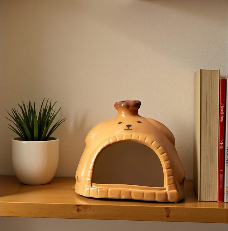 May include: A tan ceramic dog-shaped decorative mirror with a brown top and black facial features. The mirror has a rectangular opening and sits on a wooden shelf. A small potted plant and books are also on the shelf.