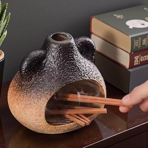 Peut inclure: Un porte-encens en céramique avec une surface texturée et un dégradé de couleurs, passant du beige clair au marron foncé. Le support a une grande ouverture et un petit trou en haut. Des bâtonnets d'encens sont visibles à l'intérieur.