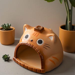 May include: A ceramic cat-shaped planter in shades of orange and tan. The planter has a large opening for plants, with a small rectangular opening on top. The cat has black eyes, whiskers, and orange stripes. A small succulent plant is visible in the background.