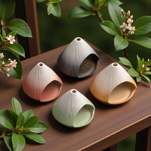 May include: Four small, cone-shaped ceramic vases with a gradient color scheme. The vases are in shades of red, gray, green, and yellow, each with a curved opening. They are arranged on a wooden surface, with greenery in the background.