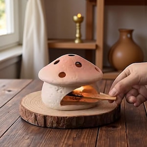 May include: A ceramic mushroom-shaped incense holder with a light pink cap and white stem, placed on a wooden slice. The holder has several holes on top and a small opening at the base. A hand is holding a wooden stick near the opening.