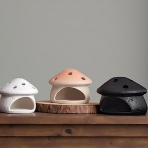 May include: Three ceramic mushroom-shaped decorative items. One is white, one is peach, and one is black. The peach mushroom sits on a wooden disc. Each mushroom has a large opening and several small holes on top.