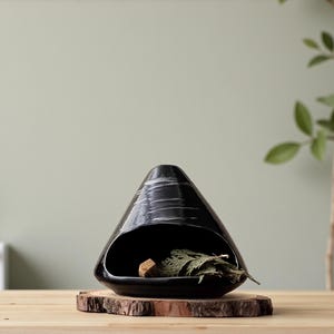 May include: A black ceramic decorative piece with a unique, cone-like shape and a large opening. The interior holds a small wooden block and dried greenery. The piece sits on a round, natural wood slice.