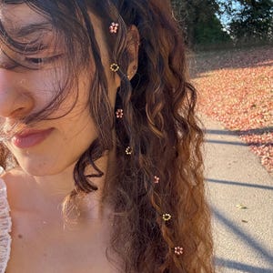 Puede incluir: Primer plano de cabello castaño rizado adornado con pequeñas horquillas florales decorativas. Los pasadores son en tonos dorados y rosas, agregando un toque de elegancia. El cabello está peinado con una onda natural, capturando la luz del sol.