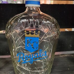 May include: A clear glass bottle with a diamond pattern, a blue and white ribbon around the neck, and a cork stopper. The bottle features a blue shield with a gold crown and the letters "KC", and the word "Royals" in blue script.