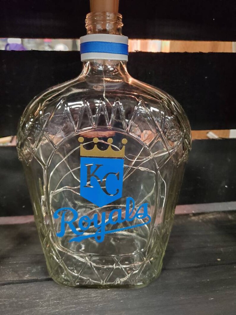 May include: A clear glass bottle with a diamond pattern, a blue and white ribbon around the neck, and a cork stopper. The bottle features a blue shield with a gold crown and the letters "KC", and the word "Royals" in blue script.