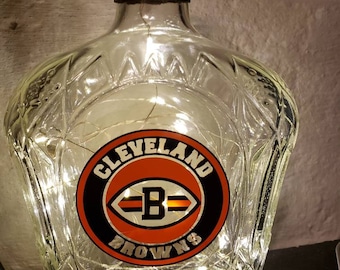 May include: A clear glass bottle with a decorative design and a cork stopper. The bottle features a Cleveland Browns logo and is illuminated with string lights. A black and red ribbon is wrapped around the neck of the bottle.