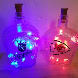 May include: Two glass bottles with cork stoppers, illuminated by string lights. One bottle features the "KC Royals" logo, the other the "KC" logo. The lights inside the bottles glow blue and red, respectively. The bottles are clear with a decorative shape.