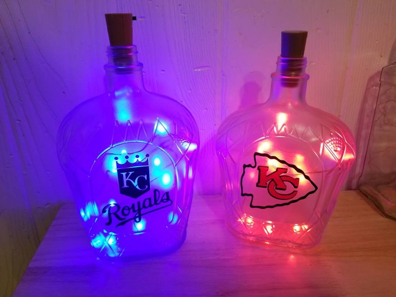May include: Two glass bottles with cork stoppers, illuminated by string lights. One bottle features the "KC Royals" logo, the other the "KC" logo. The lights inside the bottles glow blue and red, respectively. The bottles are clear with a decorative shape.