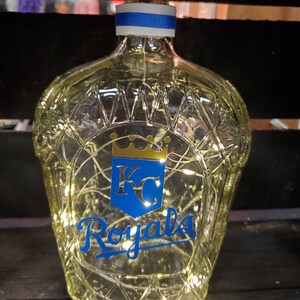 May include: A clear glass bottle with a diamond pattern, a cork stopper, and a blue and white ribbon. The bottle features the Kansas City Royals logo in blue and gold, and is illuminated with fairy lights.