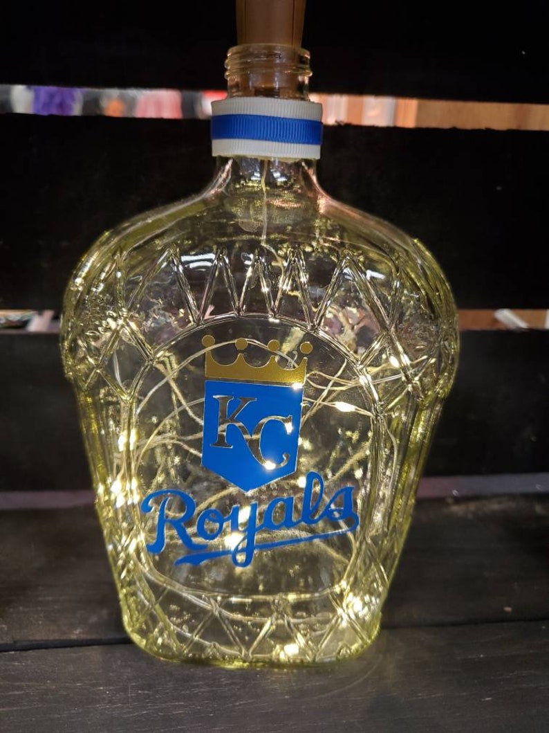 May include: A clear glass bottle with a diamond pattern, a cork stopper, and a blue and white ribbon. The bottle features the Kansas City Royals logo in blue and gold, and is illuminated with fairy lights.