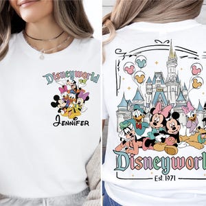 Op de afbeelding: Wit T-shirt met Disney-personages, waaronder Mickey en Minnie Mouse, Donald Duck en Goofy. De voorkant heeft een groep personages en de naam "Jennifer". De achterkant toont een kasteel en de tekst "Disneyworld Est. 1971".