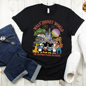 May include: Black t-shirt featuring a colorful graphic of Disney characters in front of a castle, with the text "Walt Disney World" and "The Happiest Place on Earth". The shirt is paired with blue jeans and white sneakers.