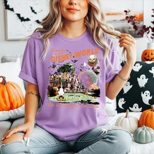 Puede incluir: Una camiseta de color morado claro con un estampado gráfico de Disney World con elementos de Halloween, incluyendo murciélagos, calabazas y un Mickey Mouse fantasmagórico. El texto dice Florida Est 1971 DISNEY WORLD y The scariest place on Earth.