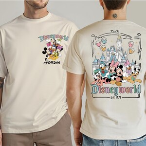 Op de afbeelding: Cr&egrave;mekleurig T-shirt met een Disney World-ontwerp. De voorkant toont Disney-personages en de naam "Jordan". De achterkant toont het Disney World-kasteel en meer personages, met "Disneyworld Est. 1971".
