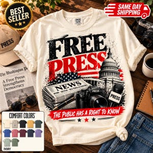 Puede incluir: Una camiseta de manga corta de color crema con un estampado gráfico con el texto FREE PRESS. El diseño incluye un periódico, una cámara, un micrófono con la etiqueta PRESS y el Capitolio de EE. UU., con el texto THE PUBLIC HAS A RIGHT TO KNOW.
