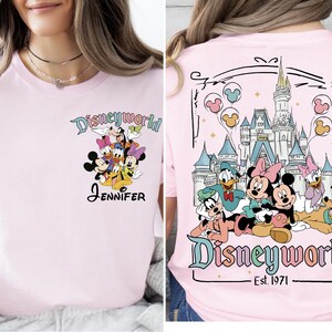 Op de afbeelding: Lichtroze T-shirt met een Disney World-ontwerp. De voorkant toont Disney-personages en de naam "Jennifer". De achterkant toont het Disney World-kasteel en meer personages, met "Disneyworld Est. 1971".