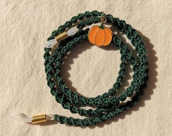 May include: A dark green macrame glasses chain featuring a small orange pumpkin charm. The cord is constructed with a knotted pattern and includes gold-toned metal coils attached to clear silicone loops at both ends for securing eyewear.