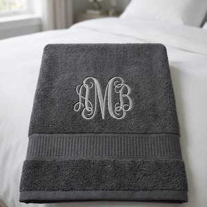 May include: A dark gray bath towel featuring a textured terry cloth surface and a horizontal ribbed band near the bottom edge. The center of the towel is embroidered with a large, white, decorative script monogram featuring the letters AMB.